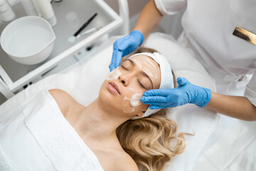 Fototapeta premium Beautician doctor in blue gloves is applying facial cleansing foam or cream on woman's face massaging skin in cosmetology clinic. Female beauty concept