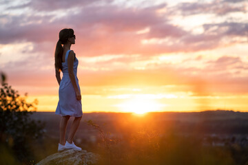 A young woman in summer dress standing outdoors enjoying view of bright yellow sunset