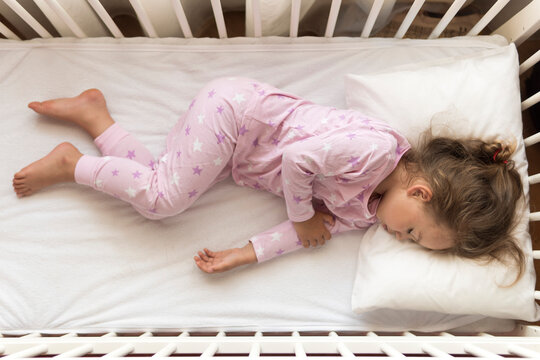 top view Cute little 3-4 years preschool baby girl kid sleeping sweetly in white crib during lunch rest time in pink pajama with pillow at home. Childhood, leisure, comfort, medicine, health concept