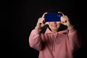 Cute young brunette in a pink hoodie uses a smartphone and laughs, communication without borders, concept portrait on a black background