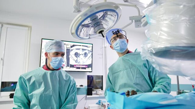 Two neurosurgeons stand over the patient on a table. Doctors apply instruments to perform operation. Professionals look at the screen.