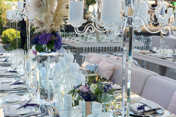 Beautiful decoration of a wedding banquet with a meal in the green garden in white colors. Very stylish setting with tropical flowers in a vase, glass candlestick set, napkins, plates, glasses cutlery