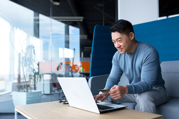 Happy Asian man shopping online, working in a modern office on a laptop, smiling and rejoicing, the chosen product, holding a credit card