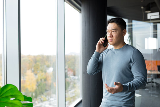 Serious and angry man talking on the phone in the office, pensive Asian at the window angry
