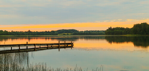 A lake with the reflection of clouds and trees at sunset. A long wooden footbridge leads into the...