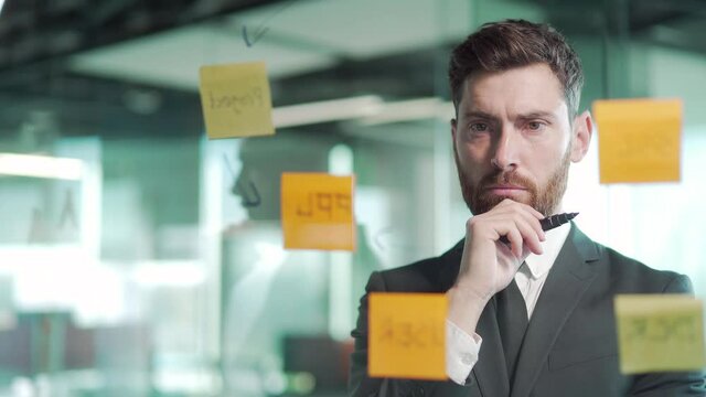 business man in modern office uses post sticky notes to share an idea. brainstorming, Bearded serious thinking male solving strategy. Use glass note wall Board and Sticky Papers. new startup posted
