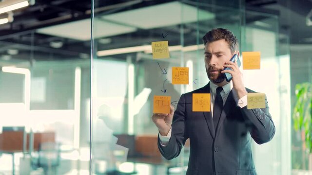 business man talking on the phone in a modern office behind a glass task partition wall with paper stacks checking information on ideas board hanging plans on colored paper stickers, plans for future