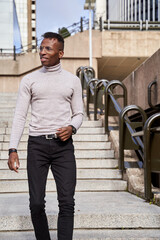 Black man in gray turtleneck and glasses looking away and walking on staircase on sunny day in city