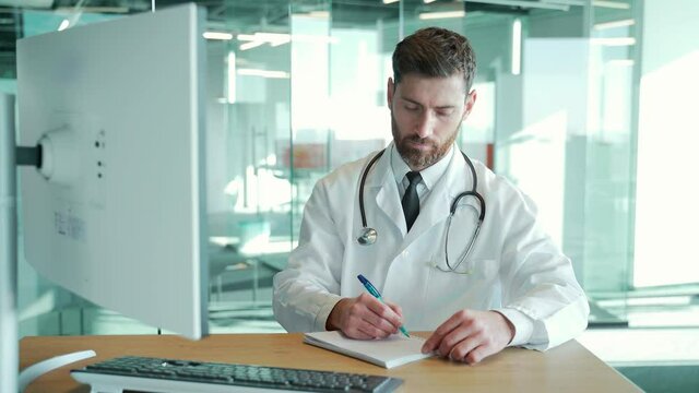 man doctor working in office at desk in modern hospital clinic. Uses pc, writes a prescription in a notebook sitting. Senior male pharmacist practitioner on computer making medical notes