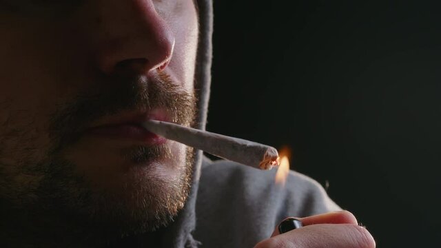 Close-up shot of a Caucasian young man&rsquo;s face with a beard and a hand, lighting rolled marijuana joint with a lighter.