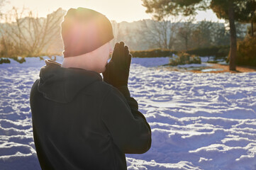 young man from behind, dressed in dark sportswear, warms up his hands while taking a break from his daily workout, in a totally snowy urban park on a sunny day during winter season