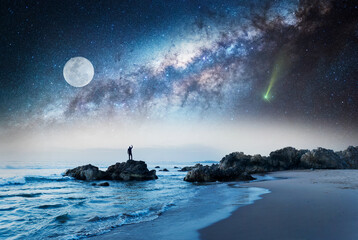 Naklejka na ściany i meble unknown person standing on the rock at the beach at night with hand up and Milky Way background 