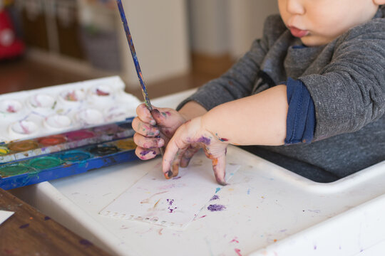 18 month old toddler watercolor painting with a brush; hands are paint covered and mouth is pursed in thought
