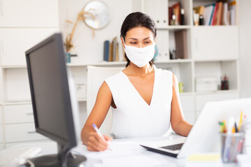 Young businesswoman in medical mask and white dress working with laptop and papers in office....