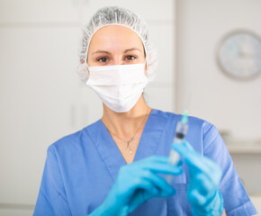 Nurse in a protective mask, working in the hospital, stands with a syringe filled with medicine in the treatment room