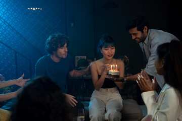 Handsome young man surprising birthday girl with a beautiful small cake with candles while surrounded with friends at midnight during party at home