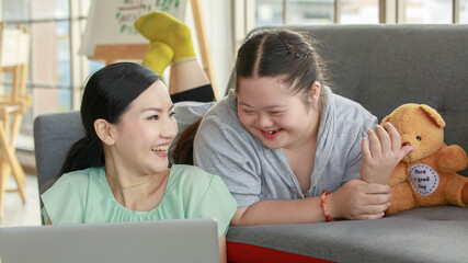 Fototapeta premium Portrait shot of Asian lovely mother sitting on floor and young chubby down syndrome autistic autism little daughter lay down on cozy sofa smiling look at camera watching cartoon from laptop computer
