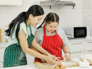 Asian lovely mother encouraging teaching young chubby down syndrome autistic autism pastry chef girl in red apron learning threshing massaging bakery flour dough with wooden rolling pin in kitchen