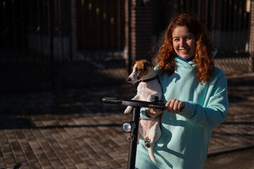 Fototapeta na wymiar A red-haired woman rides an electric scooter around the cottage village with the dog Jack Russell Terrier.