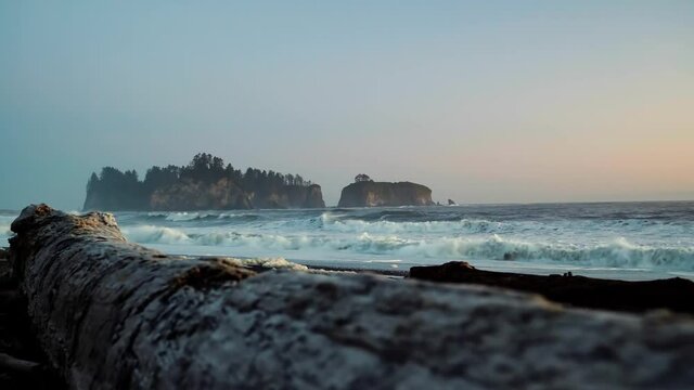 Gorgeous landscape beach shot of driftwood with waves crashing in the background, a beautiful small cliff island, and a colorful cloudy sky during sunset at the famous Ruby Beach in Forks, Washington.
