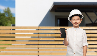 building, construction and profession concept - smiling little boy in helmet showing smartphone over living house background