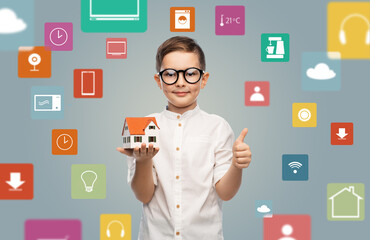 real estate, technology and people concept - smiling little boy in glasses with toy house model showing thumbs up over virtual icons on grey background