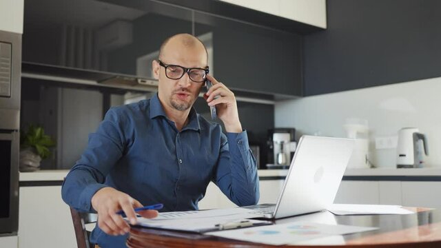 Male employee with eyeglasses wearing shirt working from home, talking on phone and checking papers, using laptop, kitchen devices on blurred background. Concept of remote work and outsource