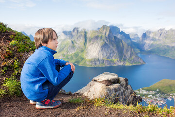 Cute child, standing on top of the mountains and looking down on Reine after climbing Reinebringen treeking path with lots of stairs, Lofoten, Norway