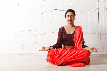 Young adult indian woman in sari meditating yoga lotus pose zen like with ok sign mudra gesture at...