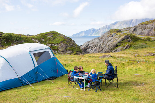 Happy family with three kids, wild camping in Norway summertime, people having breakfast adn coffee on a cliff next to a fjord