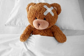 medicine, healthcare and childhood concept - ill teddy bear toy with medical patch on head lying in bed