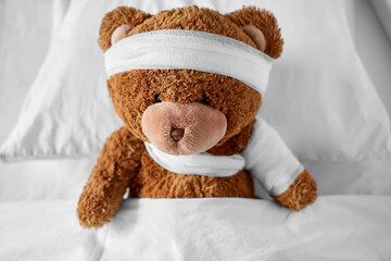 medicine, healthcare and childhood concept - ill teddy bear toy with bandaged paw and head lying in bed