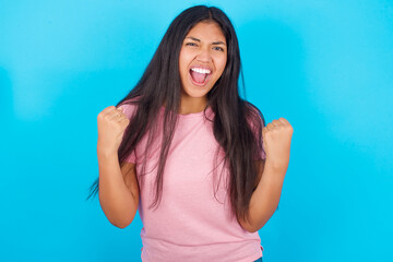 Fototapeta na wymiar Portrait of Young hispanic girl wearing pink T-shirt over blue background looks with excitement at camera, keeps hands raised over head, notices something unexpected reacts on sudden news.