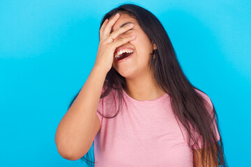 Charismatic carefree joyful Young hispanic girl wearing pink T-shirt over blue background  likes laugh out loud not hiding emotions giggling hear funny hilarious joke chuckling facepalm.