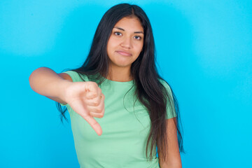 Young hispanic girl wearing green T-shirt over blue background looking unhappy and angry showing...