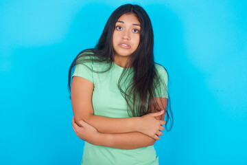Fototapeta na wymiar Young hispanic girl wearing green T-shirt over blue background shaking and freezing for winter cold with sad and shock expression on face.