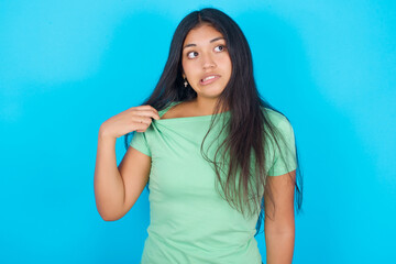 Fototapeta na wymiar Young hispanic girl wearing green T-shirt over blue background stressed, anxious, tired and frustrated, pulling shirt neck, looking frustrated with problem