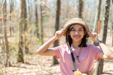 Happy beautiful young adventurous woman is smiling, touching her hat and looking at camera in dry forest while walking and hiking to epic mountain with copy space. Outdoor adventure concept.