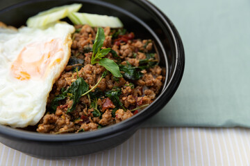 Closeup of Thai traditional food, Flavorful appetizing food as Spicy fried pork with basil leaves, fried egg and cucumber in black modern bowl on clean minimal cream and green table cloth background