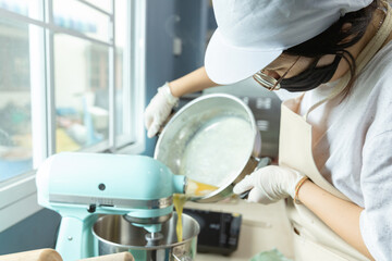 Happy young Asian woman with protective face mask is bringing bakery material to bakery mixer to bake homemade cake or bakery first time, learning new skills when home quarantine, Coronavirus spread.