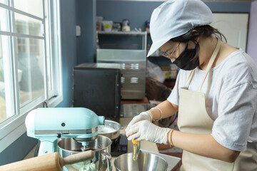 Happy young Asian woman with protective face mask, apron is smiling, cracking eggs, preparing bakery material to bake homemade cake or bakery first time, learning new skills when home quarantine.