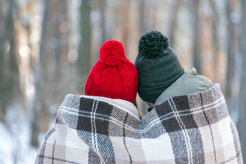 Guy and girl in knitted hats with buboes in winter in park are covered with warm blanket. Back view