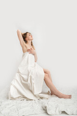 Fototapeta na wymiar Young caucasian dreaming woman is sitting covered herself with white sheet and poses. Concept of good morning. White background. Vertical frame