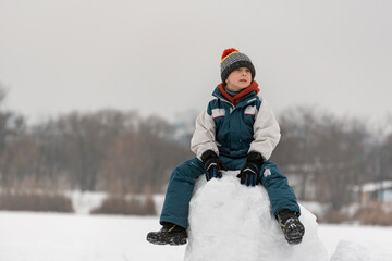 Fototapeta na wymiar Smiling boy of seven years old sits on snow tower. Child builds snow castle. Winter time.