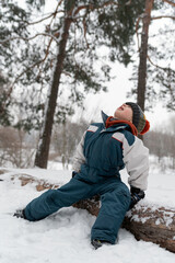 Fototapeta na wymiar Portrait of boy in the winter forest. Child sits on log surrounded by snow, raising his head to the sky. Side view. Vertical frame