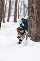 Cheerful boy peeks out from behind tree. Child in knitted hat walks through winter snow-kept park. Vertical frame