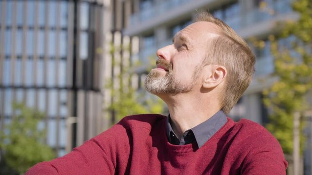 A middle-aged handsome Caucasian man looks around as he sits in a street in an urban area - office buildings in the blurry background - closeup