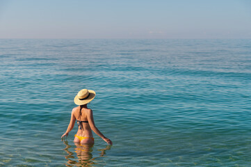 Back view of a slender caucasian young woman standing waist-deep in calm transparent sea water in a swimsuit and a straw hat