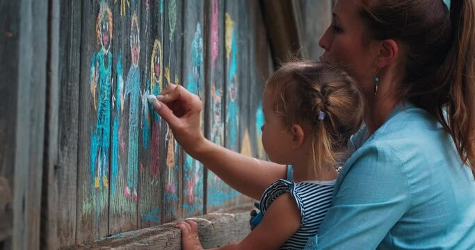 Garden art. Mother and daughter draw with colourful chalks on the wooden fence in the garden