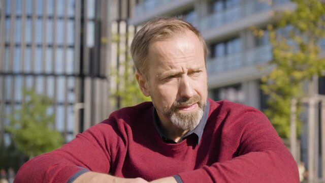 A middle-aged handsome Caucasian man thinks about something as he sits in a street in an urban area - office buildings in the blurry background - closeup